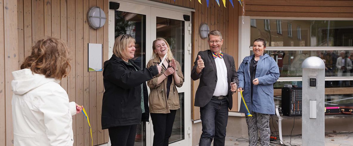 Fem personer vid entré pyntad med blågula vimplar. Tre personer har en sax i sin hand och skrattar, klappar och gör tummen upp.