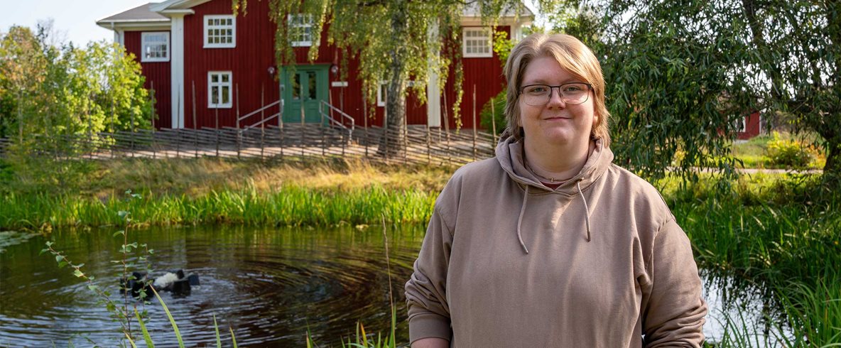 Kvinna i kort rakt brunt hår, glasögon och brun huvtröja ler mot kameran med lummig grönska, damm och rött hus i bakgrunden.