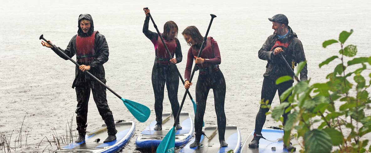 Fyra personer paddlar ståendes i en sjö, i regn.