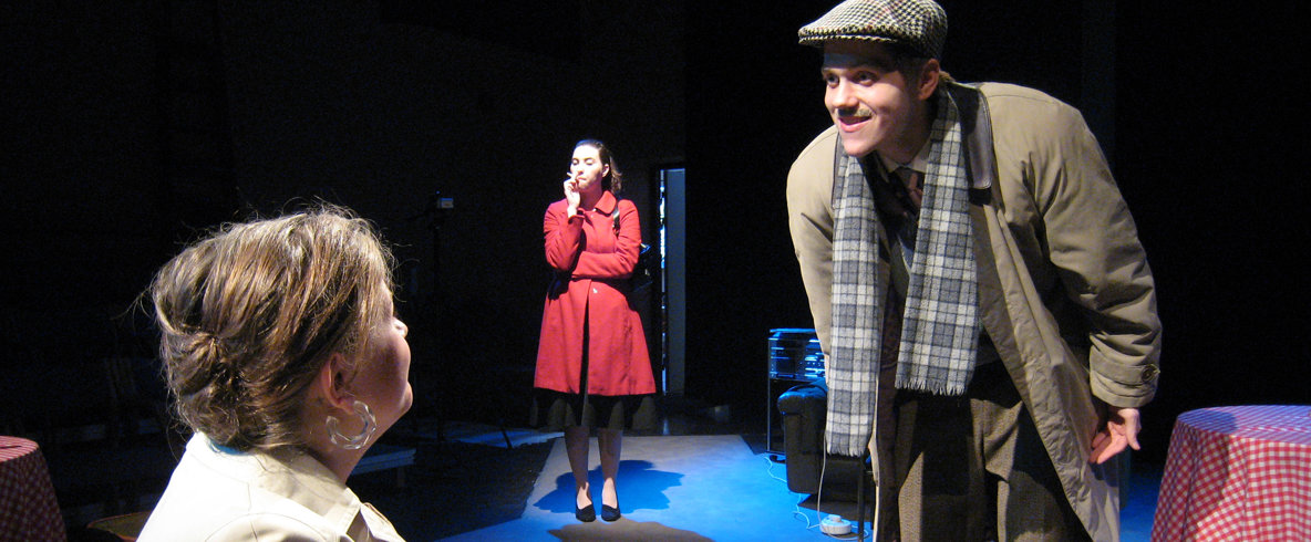 En man böjer sig ner och pratar med en kvinna som sitter ner medan en kvinna står och röker i bakgrunden.