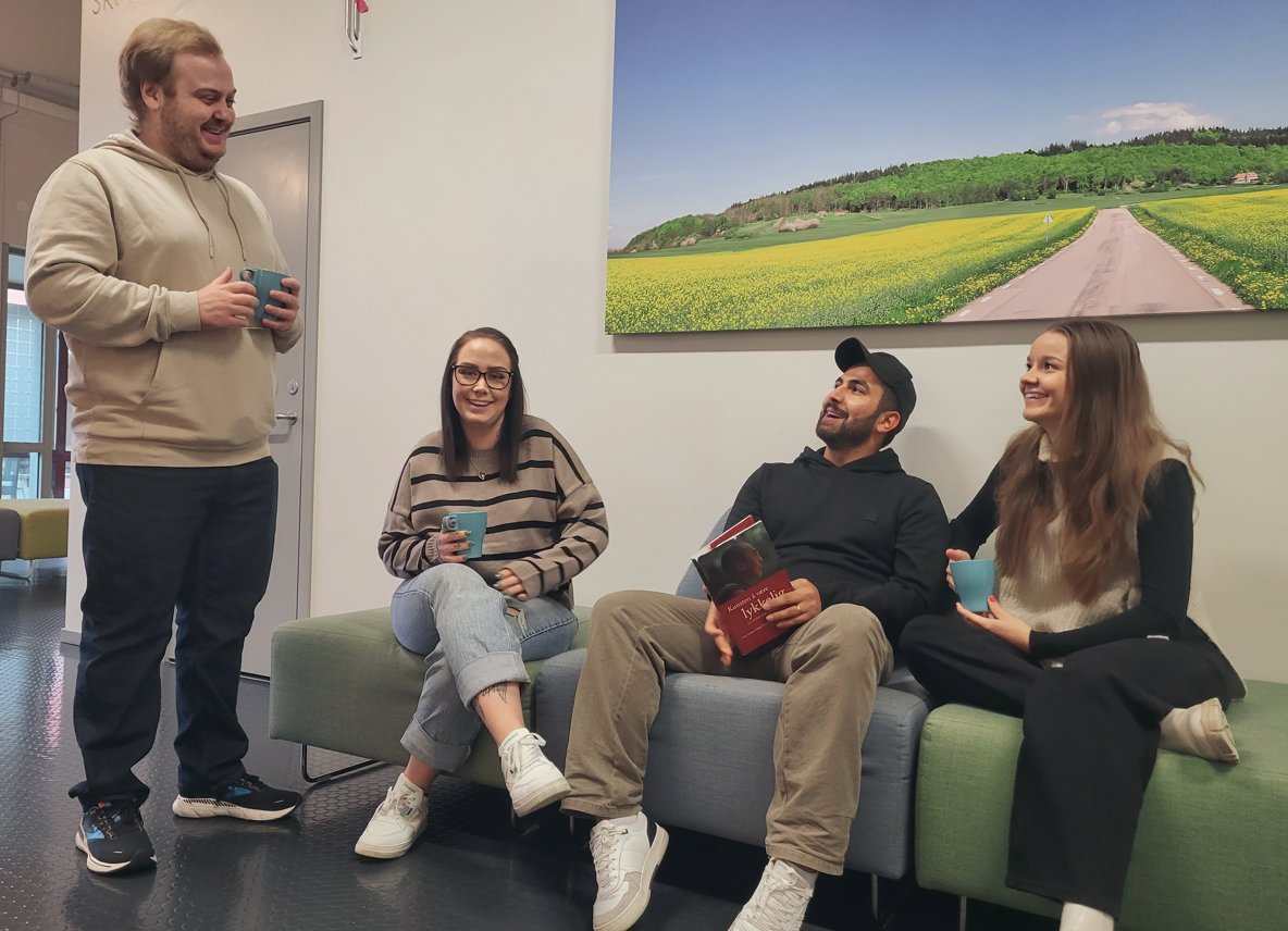 Fyra personer i en korridor. Tre sitter på en soffa och tittar upp på en fjärde. Alla skrattar åt något den fjärde precis sagt.